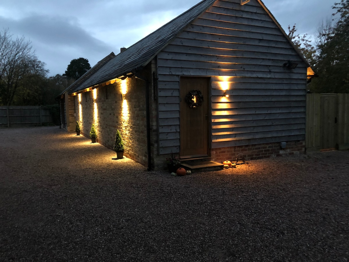 The exterior of the barn is illuminated by warm lighting, highlighting the wooden siding and entrance door. Decorative plants and a subtle pathway lead to the door, creating a welcoming ambiance in the evening light.