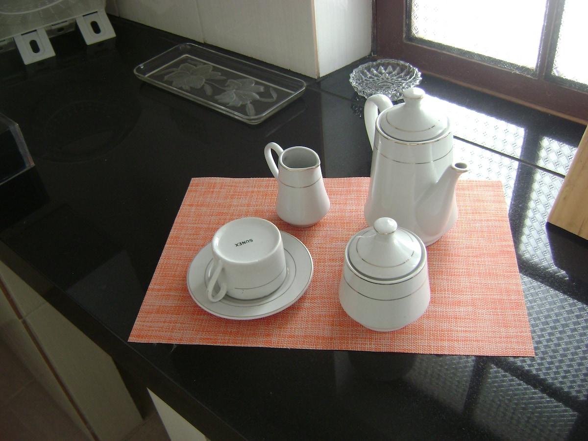 A white teapot, cream jug, and sugar bowl are arranged on a textured orange placemat. A cup and saucer sit beside them, all featuring simple silver stripes. Natural light filters through a nearby window, illuminating the polished black countertop.
