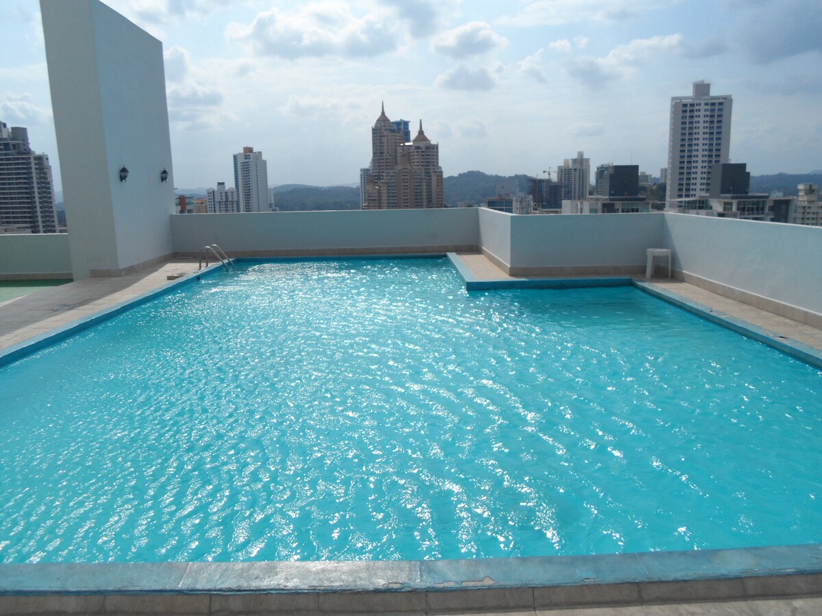 A spacious rooftop swimming pool reflects the sky, surrounded by a non-slip deck. Skyscrapers and distant hills create an urban backdrop, emphasizing the scenic elevation of the location. The pool is designed for relaxation, offering a tranquil space for guests to unwind on sunny days.