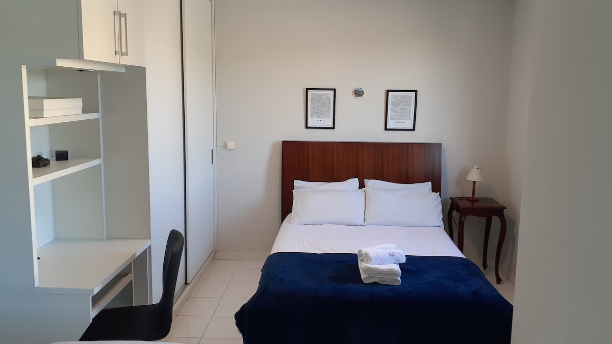 A well-appointed sleeping area features a spacious bed covered with a deep blue blanket and neatly arranged white towels. A small desk and chair sit nearby, complemented by a minimalist side table with a lamp. Artwork adorns the walls, contributing to the room's aesthetic.