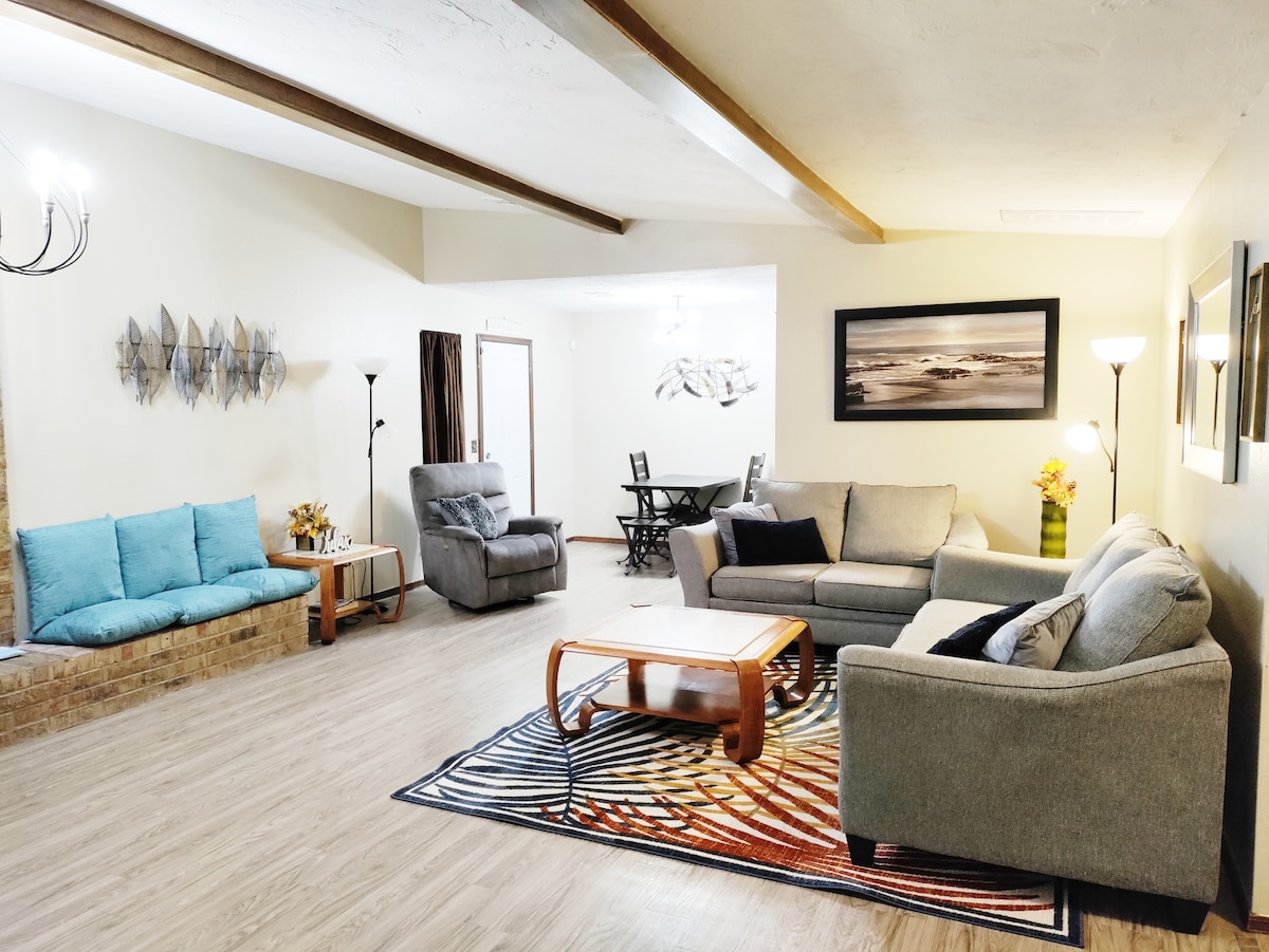 A spacious living area features a comfortable grey sofa, armchairs, and a coffee table on a large area rug. Natural light illuminates the space, highlighting the wooden beams overhead. A dining table is placed in the background, accompanied by a wall-mounted decorative piece.