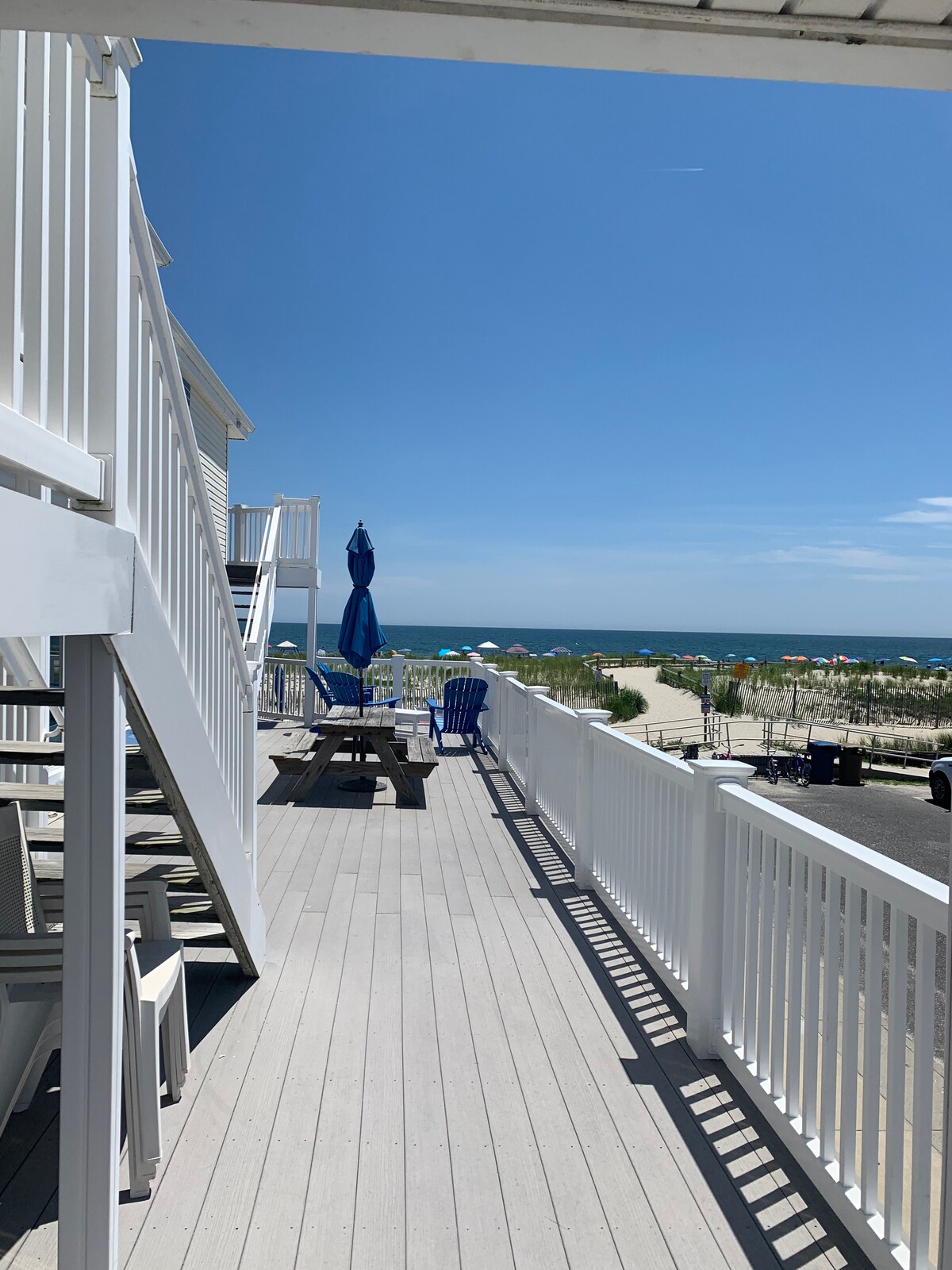 A spacious deck is shown with a wooden dining table and chairs, inviting relaxation. A large umbrella provides shade, while the ocean is visible in the distance. Beach umbrellas can be seen dotting the sandy shore under a clear blue sky.