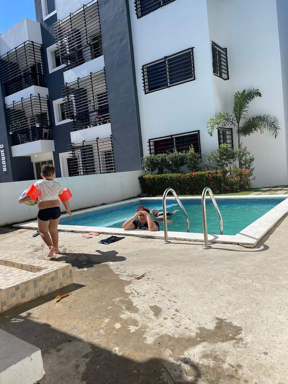 A swimming pool is seen in a sunny outdoor area, surrounded by a minimalistic concrete deck. People are present, one is immersed in the pool while another holds colorful pool accessories. The backdrop features a modern multi-story building with several windows.