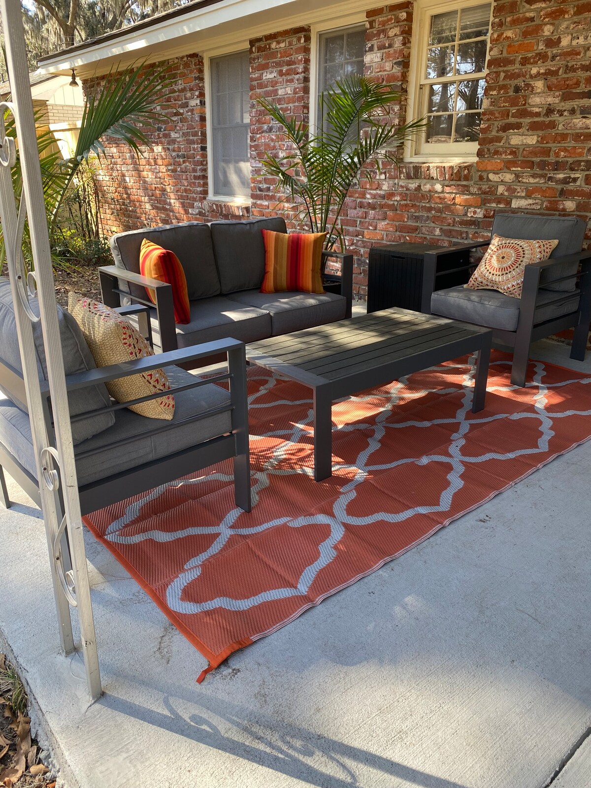 An outdoor seating area features a gray sectional and chairs with colorful cushions, arranged around a low table. A patterned orange rug sits beneath the furniture, while potted plants enhance the space against a backdrop of exposed brick.