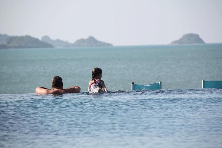 An infinity pool extends towards the horizon, blending seamlessly with the Gulf of Thailand. Two individuals relax on the pool's edge, enjoying the tranquil views. Hills are visible in the distance, while the calm water reflects the clear sky.