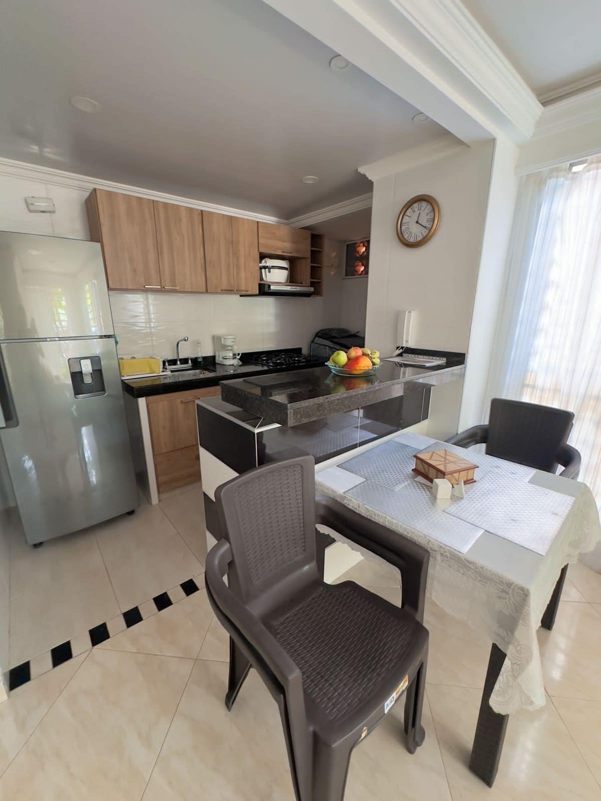 The kitchen area features modern cabinetry with ample storage and is equipped with a stove, refrigerator, and microwave. A small dining table is set for two, accompanied by two plastic chairs. Natural light enters through the nearby window, enhancing the space.