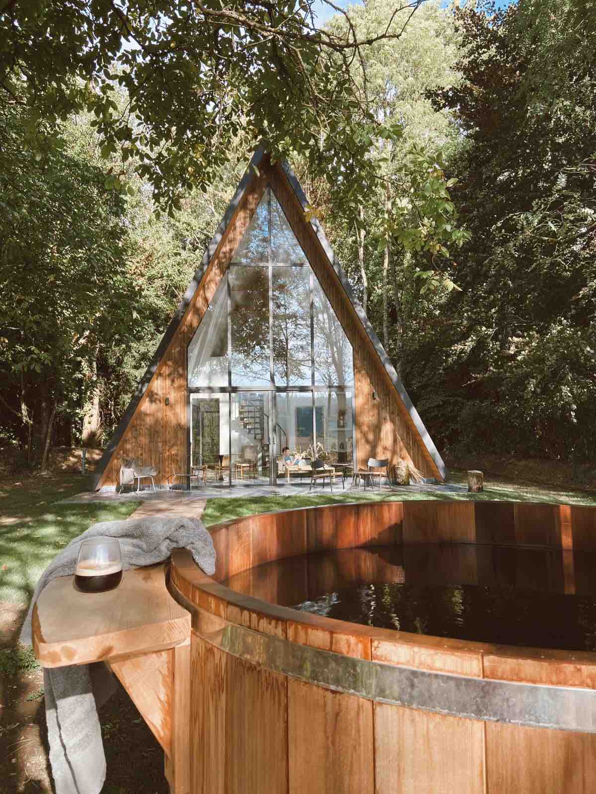 A triangular wood and glass lodge is nestled among trees, featuring large glass walls that invite natural light. A private wooden hot tub is positioned at the forefront, offering an inviting space for relaxation surrounded by nature.
