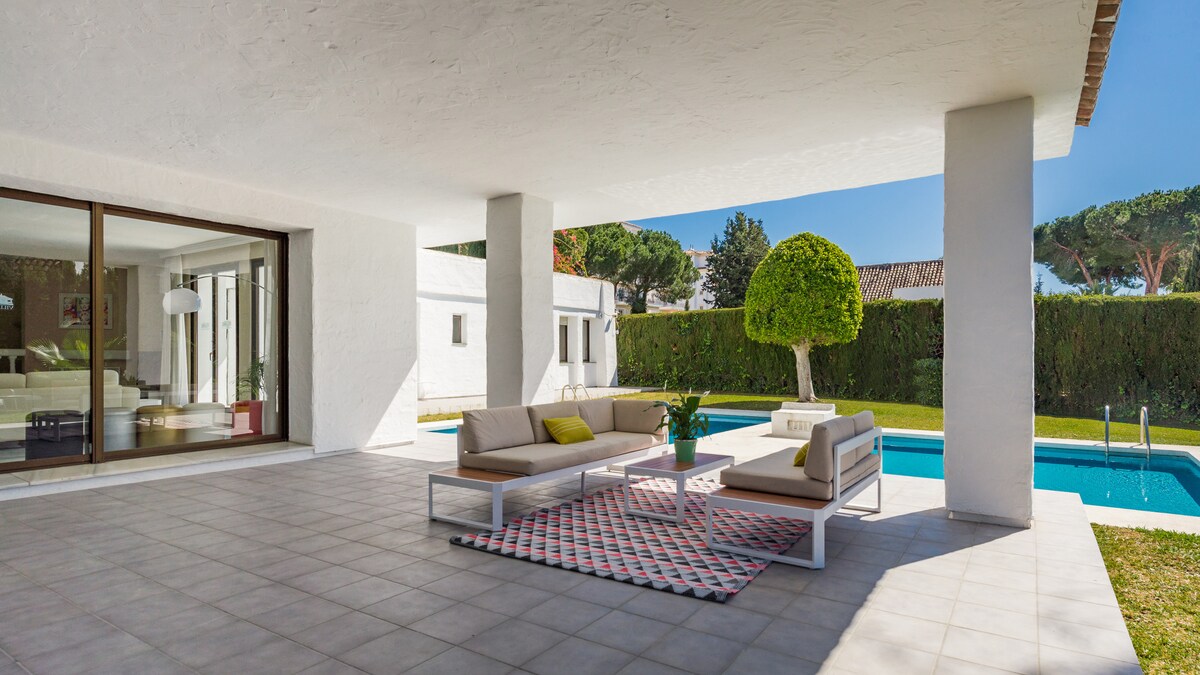 A spacious outdoor terrace features modern seating arranged around a central coffee table. A geometric area rug adds color underfoot, complementing the clear blue waters of the pool visible just beyond. Lush greenery provides privacy, enhancing the serene outdoor atmosphere.