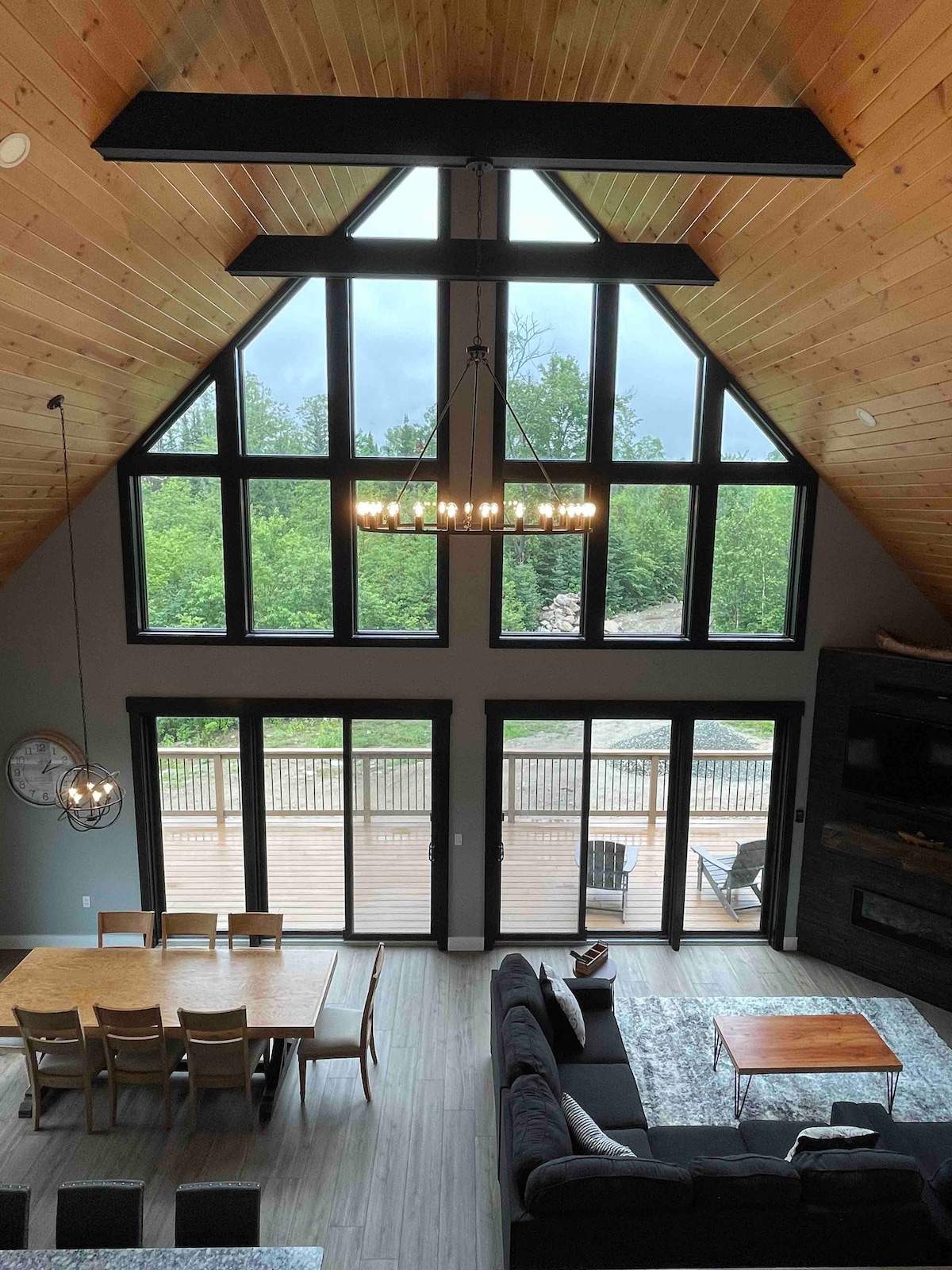 An open living space features a high wooden ceiling, accentuated by a modern chandelier. Large windows frame scenic views of greenery, allowing natural light to fill the room. A wooden dining table is arranged with chairs, and a comfortable seating area is visible.