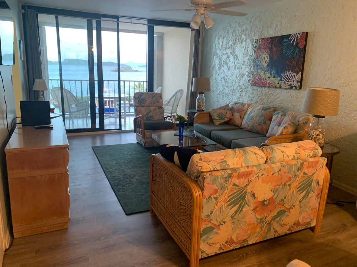 A cozy living area features a blend of rattan and upholstered furniture, including a spacious sofa and two matching chairs. A decorative rug lies underfoot, and a flat-screen TV is positioned on a wooden dresser. Large sliding glass doors open to a balcony with ocean views.