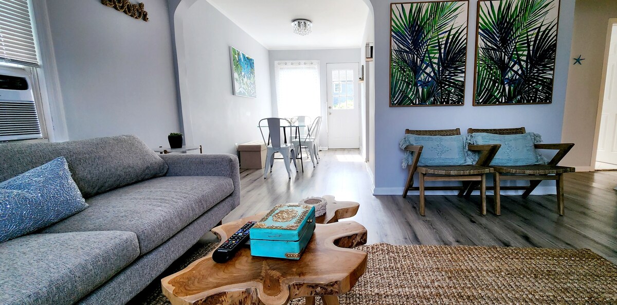 A cozy living area is highlighted by a large gray sofa and a unique wooden coffee table. Two framed tropical prints adorn the wall, while a light-filled dining area with metal chairs is visible in the background. A woven rug adds a natural touch to the space.