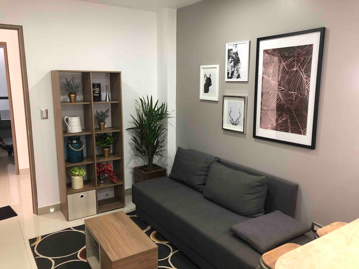 A modern living area features a gray sofa against a neutral wall. Framed art pieces are displayed above the sofa, and a shelf filled with plants and decorative items is located alongside it. A patterned rug adds warmth to the space.