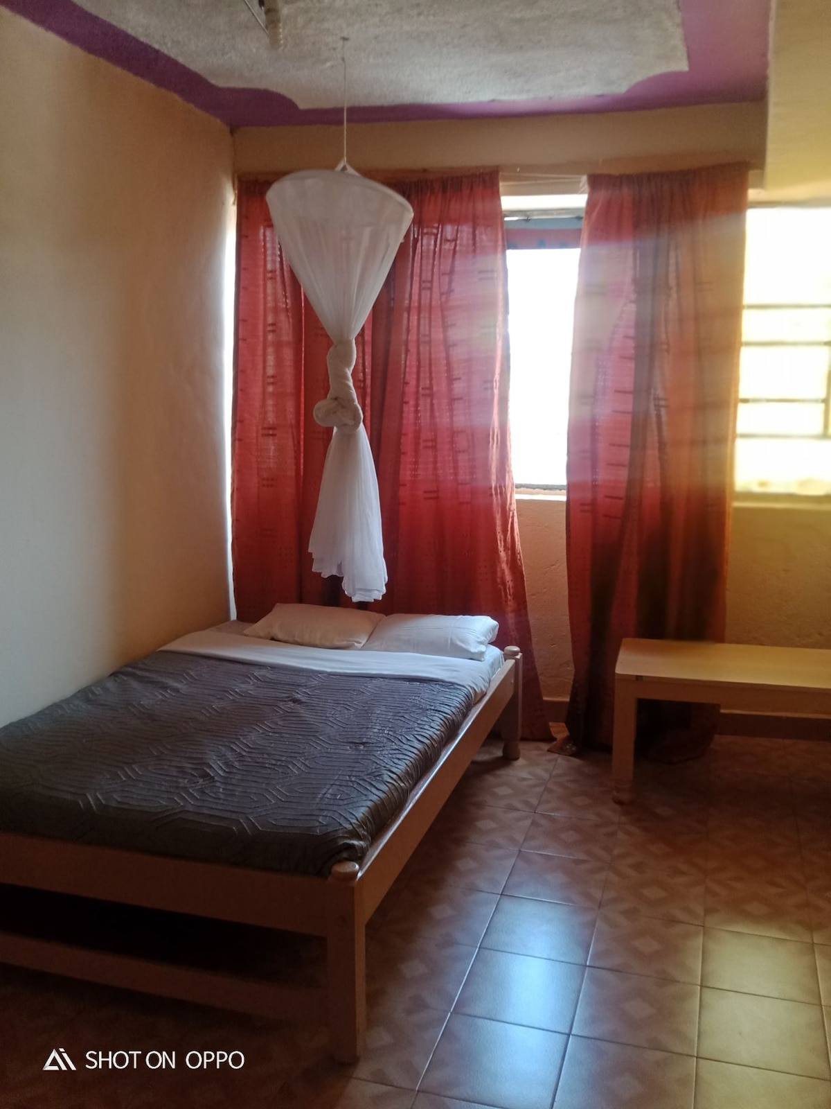 A simple bedroom features a single bed with a gray patterned mattress and white bedding. Sheer white curtains frame the window, allowing natural light to filter through. A wooden bench is positioned against the wall, complementing the warm-toned walls and tiled floor.