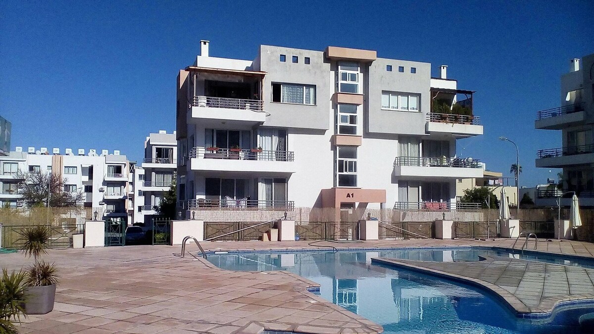The apartment complex features a modern design with multiple levels and balconies. A swimming pool is visible in the foreground, surrounded by a tiled deck. Sun loungers and small plants provide elements of leisure, while a clear blue sky creates a bright atmosphere.