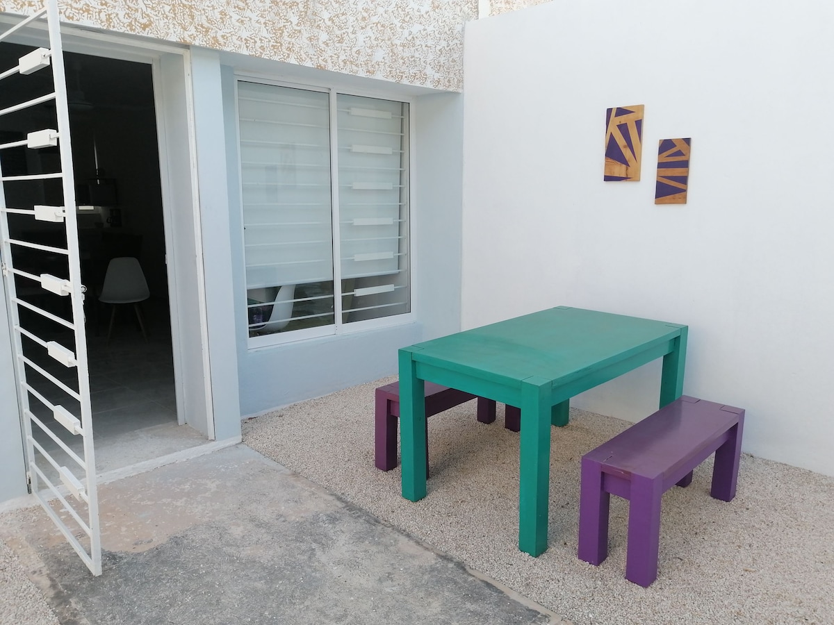 An outdoor seating area is present, featuring a colorful turquoise table surrounded by purple benches. The space is designed with light gravel underfoot, and a large window offers a view of the interior. Two abstract artworks adorn the wall.