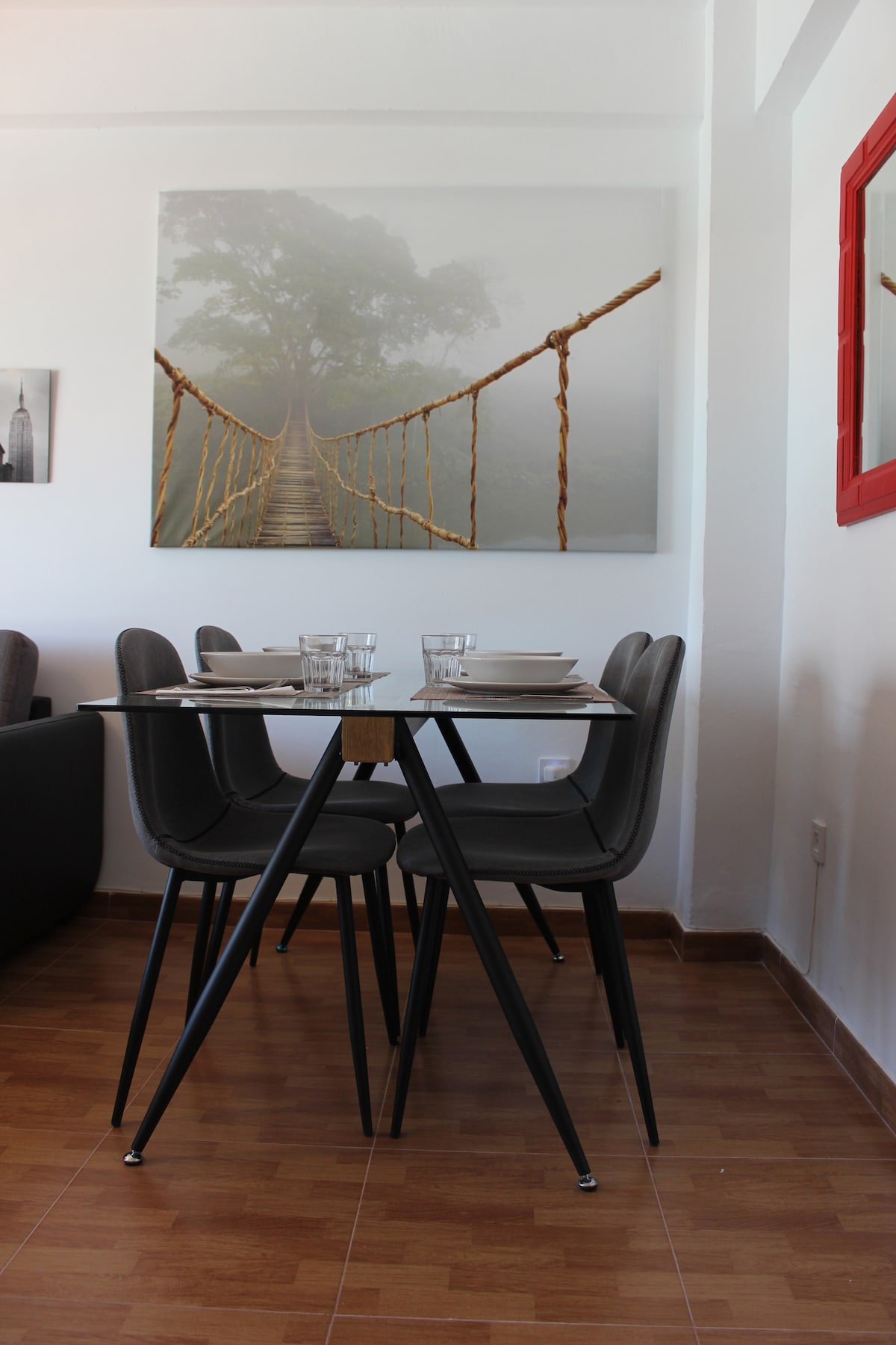 A dining area is visible with a modern table set for four, featuring glassware. A large artwork depicting a foggy bridge scene is displayed on the wall, complementing the contemporary atmosphere. The floor is covered in light-colored tiles, enhancing the bright and open space.