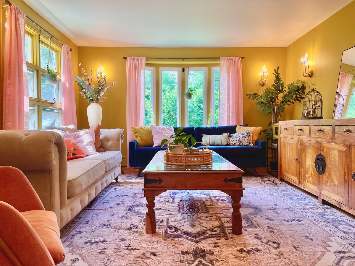 A cozy living area features a neutral sofa and a deep blue couch, complemented by soft pink curtains. A vintage-style wooden coffee table sits atop an intricately patterned area rug. Green plants and decorative accents enhance the inviting ambiance, with natural light filtering through the large windows.