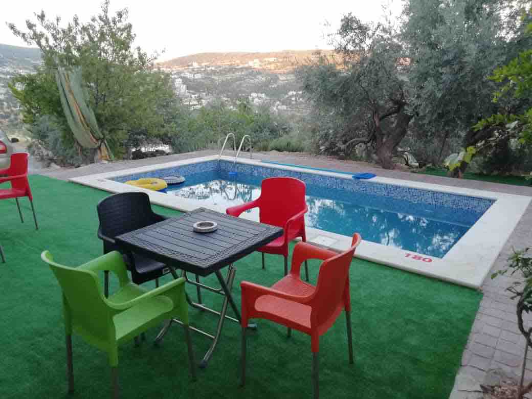 A private pool area is surrounded by lush greenery, featuring a rectangular pool with a light blue tile border. A small table with four colorful chairs is set nearby, offering a relaxing space for outdoor gatherings.