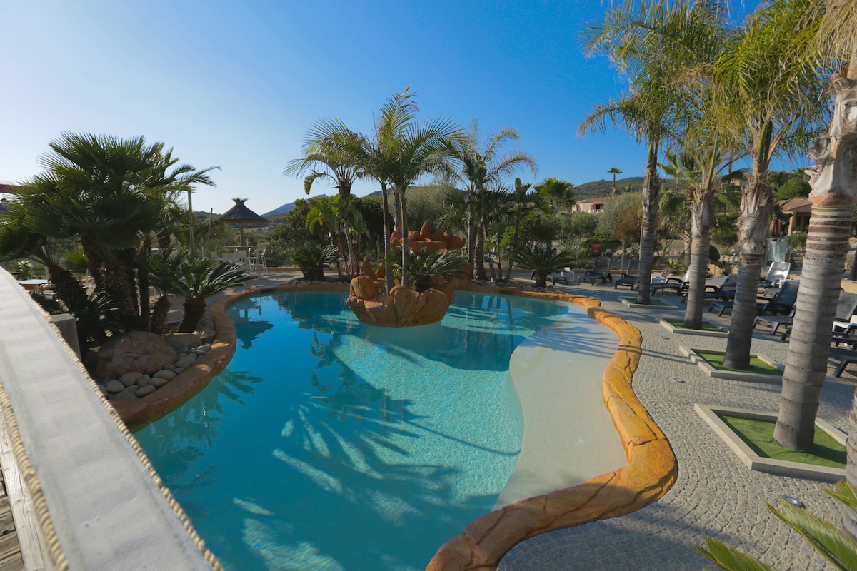A serene pool area is showcased, featuring a winding shape surrounded by lush greenery and palm trees. Comfortable lounge chairs are arranged nearby for relaxation. The clear water reflects the blue sky, creating a tranquil atmosphere for guests to enjoy.