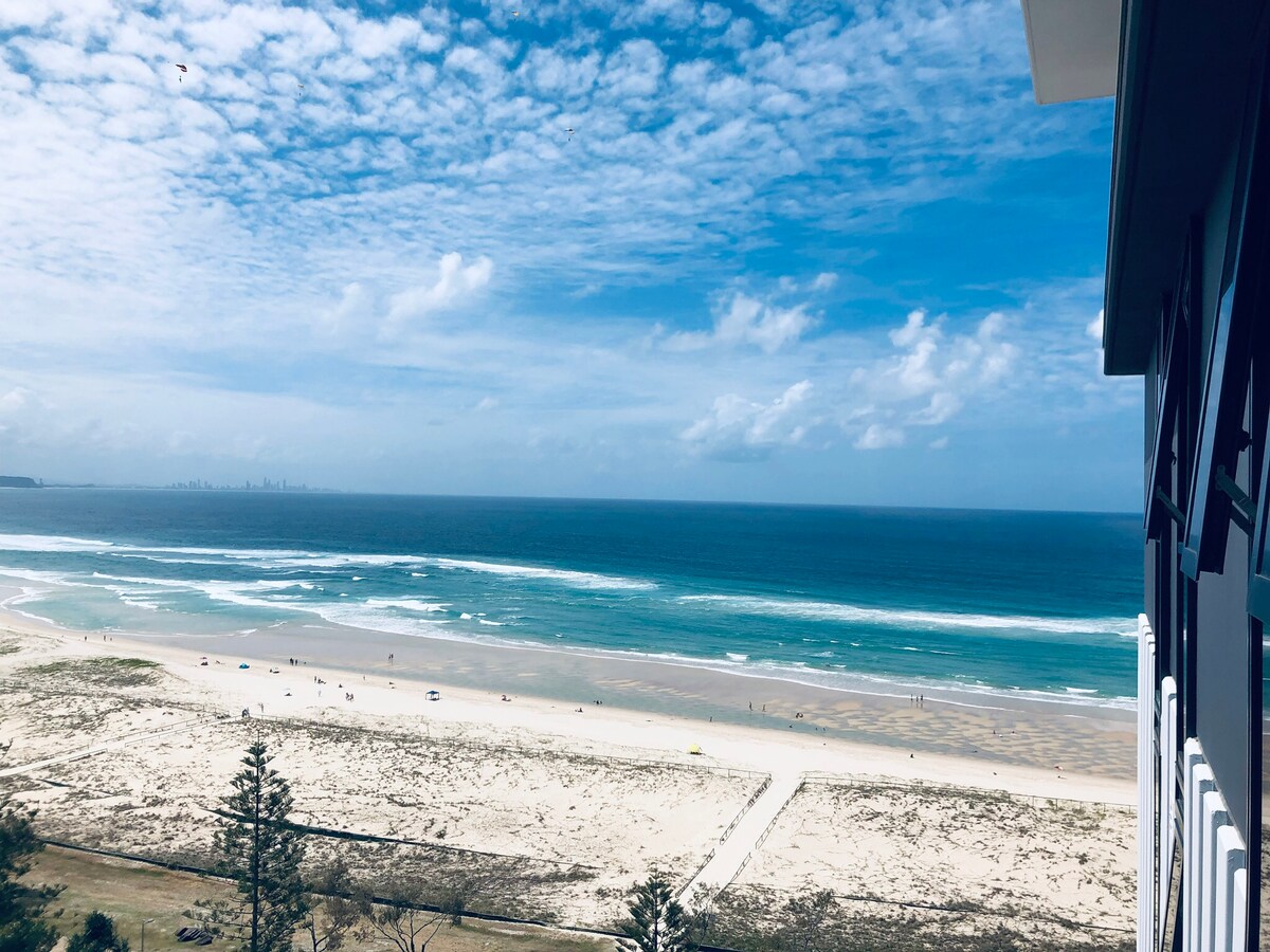 A sweeping view of the coastline extends from the apartment, featuring soft sand and rolling waves. The vibrant blue ocean contrasts with the cloud-covered sky, while distant figures are seen enjoying the beach. The Surfers Paradise skyline is visible along the horizon.
