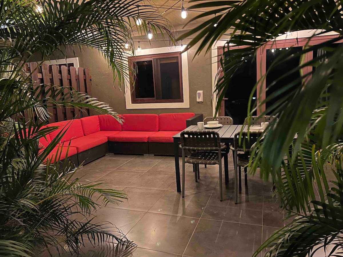 An outdoor seating area is surrounded by lush greenery. A long red sectional sofa is positioned against the wall, complemented by a dining table with several chairs. Soft lighting creates a gentle ambiance, enhancing the space's privacy and comfort.