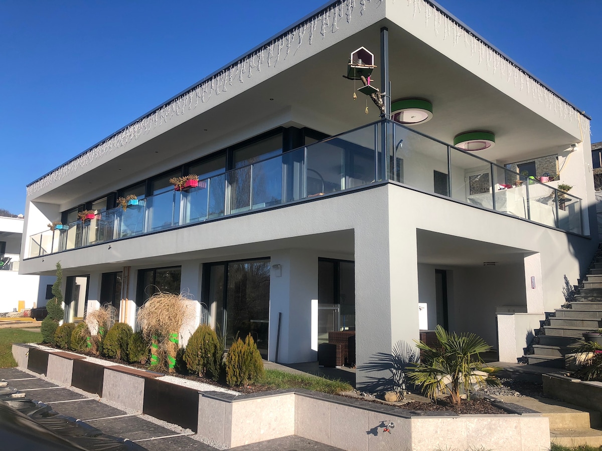 The modern building features a spacious balcony with glass railing and decorative planters. The exterior is designed with clean lines, and greenery is thoughtfully integrated into the landscape. A staircase leads to the entrance, while a well-maintained garden area enhances the overall appeal.