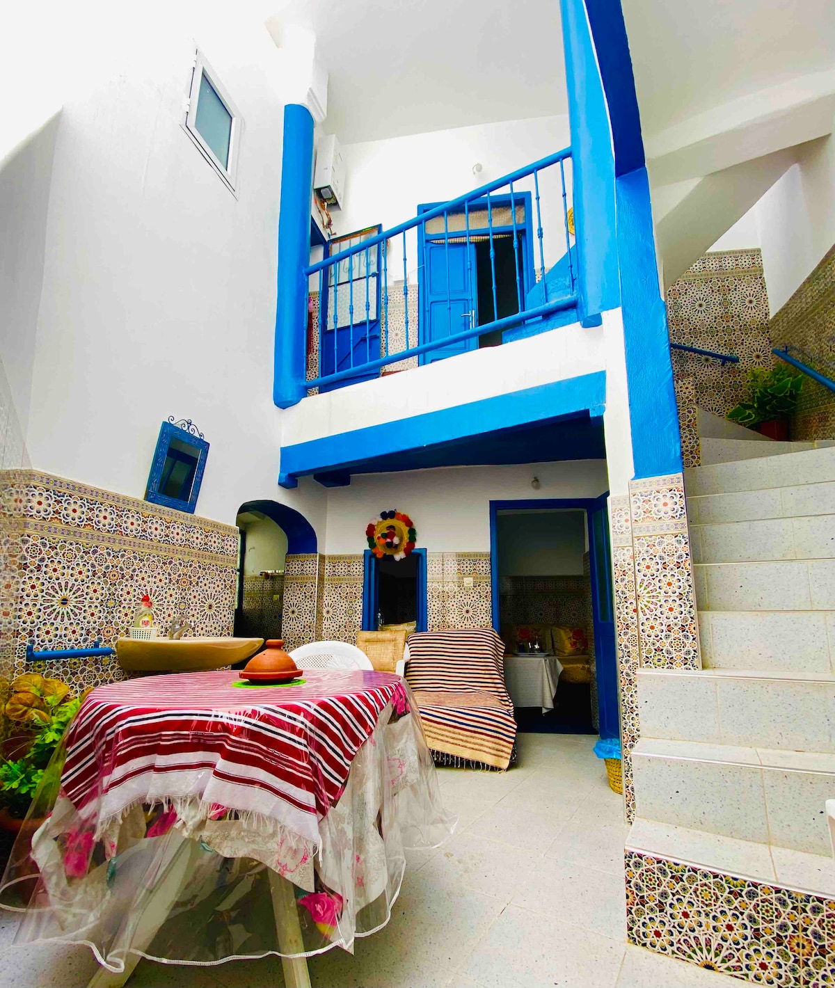 An inviting courtyard is present, featuring tiled walls with intricate patterns and a table topped with a decorative cloth. A staircase leads to the upper level, while contrasting blue accents frame the space. A glimpse of surrounding rooms can also be seen.