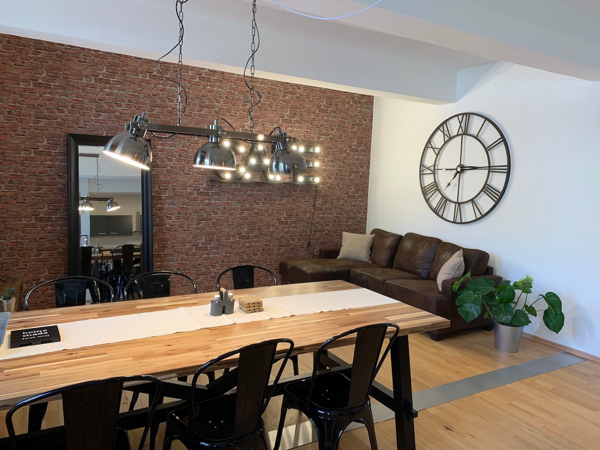 A dining area features a long wooden table with black chairs, paired with a decorative runner. Industrial-style lighting hangs above. A large clock adorns the wall next to a cozy brown sofa, and a mirror reflects the space, enhancing the open feel.