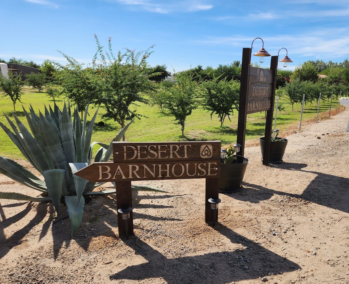 A wooden signpost displaying 'Desert Barnhouse' is positioned prominently, guiding visitors along a gravel path. Lush greenery is visible on either side, complemented by decorative pots and a large agave plant, enhancing the inviting entrance to the property.