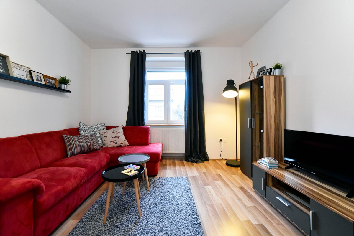 A bright living area features a red sectional sofa adorned with patterned cushions. A round black coffee table sits on a textured rug, while a tall lamp provides soft lighting. A window with light curtains allows natural light to enter, complemented by a shelf displaying framed photos.