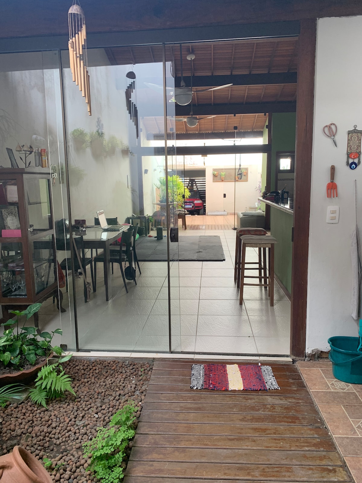 A spacious entryway features large glass doors, providing a view into a bright and airy open space. A wooden floor leads in, complemented by decorative plants and a small rug. The dining area, furnished with a black table and chairs, can be seen in the distance.