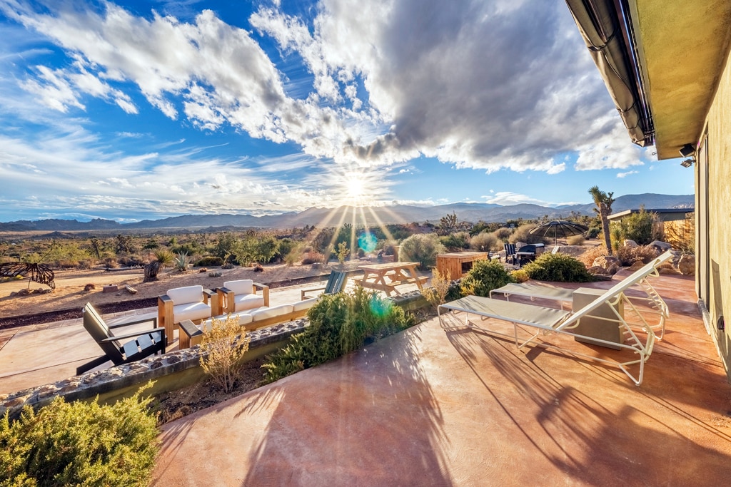 A sunlit patio area is highlighted, featuring lounge chairs and a wooden dining table. Rolling desert landscapes and distant mountains are visible, with a vibrant sky displaying the sun breaking through clouds. Lush landscaping surrounds the space, enhancing the outdoor experience.