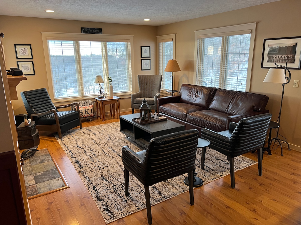 A welcoming living room is presented, featuring a large leather sofa, two patterned armchairs, and a coffee table atop a textured rug. Natural light filters through multiple windows, illuminating the space and creating a cozy atmosphere perfect for gathering.