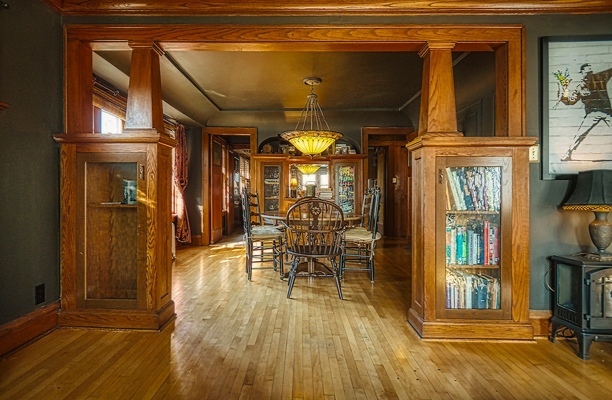 A warm dining area features a wooden table surrounded by six chairs, illuminated by an elegant chandelier. The space is framed by tall wooden pillars, with built-in shelves displaying various books. Natural light streams through large windows, highlighting the polished hardwood floor.