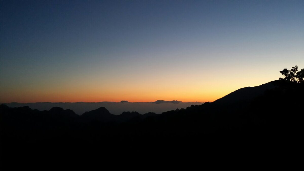A serene view of a mountain landscape features a gradient of colors from deep blue to orange at sunset. Silhouettes of distant peaks and rolling hills provide a sense of depth, while the sky transitions softly into twilight.