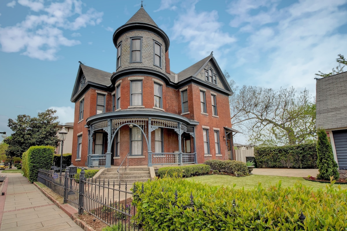 A historic brick building features a prominent turret and intricately detailed trim. The inviting front porch is supported by decorative columns, surrounded by manicured green hedges and well-kept landscaping. A pathway leads to the entrance, providing a welcoming connection to the surroundings.