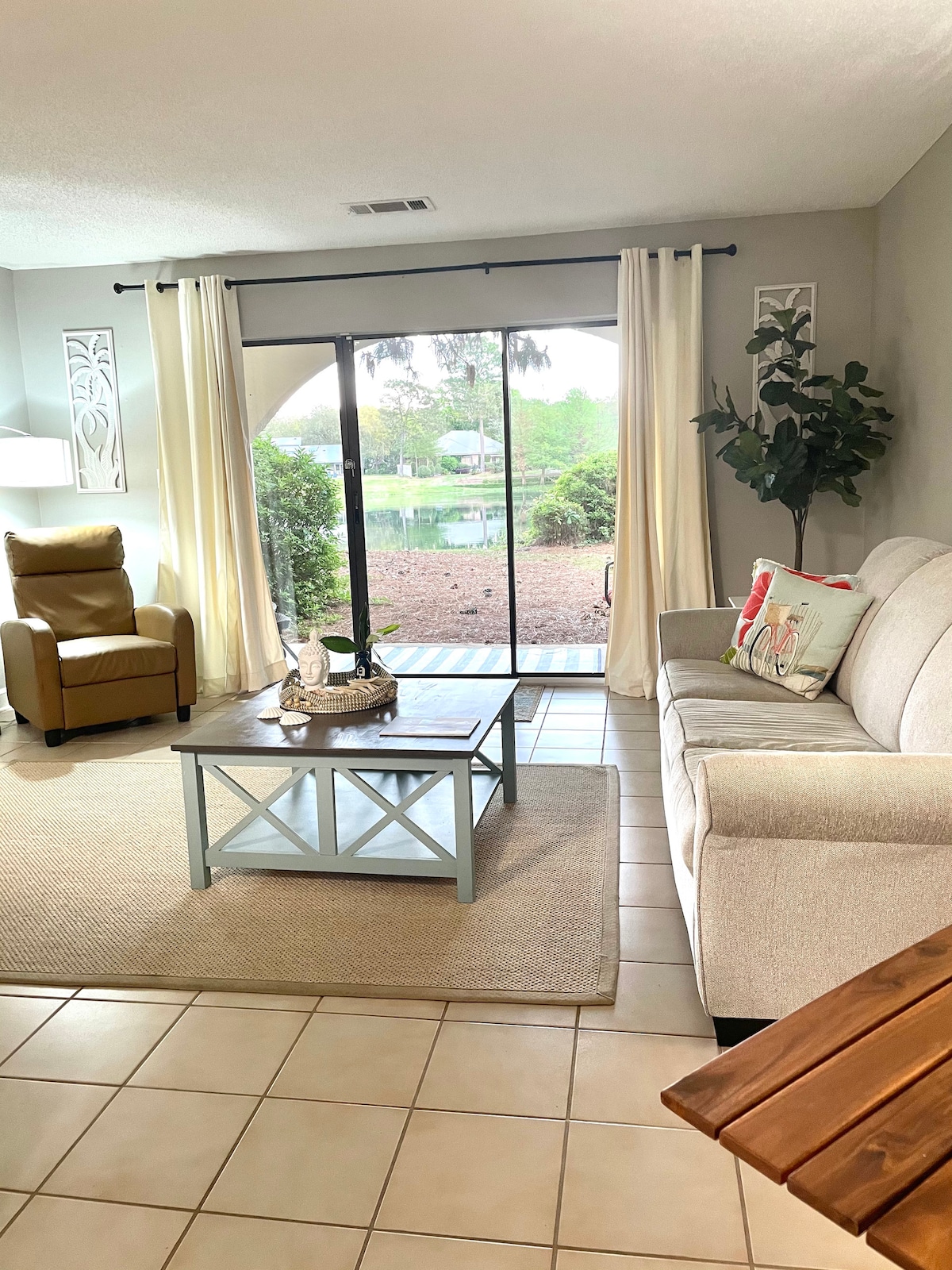 A bright living area features a comfortable light-colored sofa paired with a brown recliner. A coffee table sits in the center on a neutral area rug, with sheer curtains framing the large sliding glass door that opens to a serene lake view.