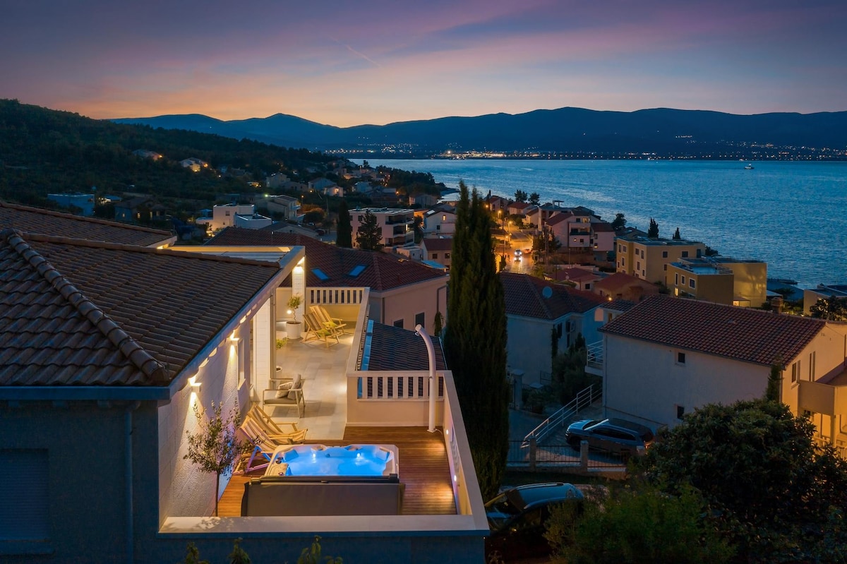 An aerial view reveals a modern penthouse terrace featuring a private jacuzzi, surrounded by sunbeds. The setting sun casts a warm glow over the tranquil sea and distant hills, while city lights begin to twinkle along the coastline.