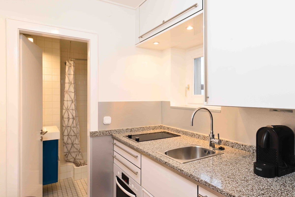 A modern kitchen is showcased with sleek cabinetry and a spacious countertop made of patterned granite. Appliances include a stovetop, oven, and a coffee maker. A doorway leads to a bathroom area, where a shower curtain and tiled floor are visible.