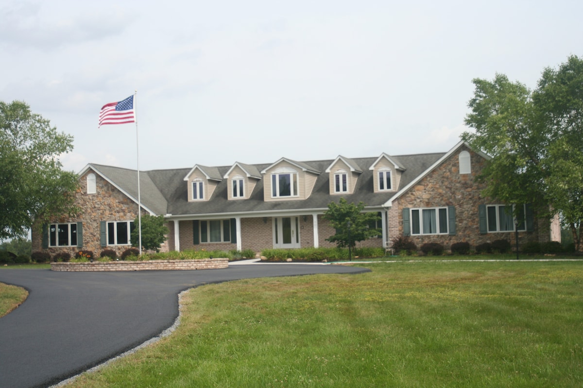 The exterior of a spacious brick and stone house is shown, featuring multiple gabled windows and a prominent American flag. A circular driveway leads to a landscaped front yard, surrounded by lush green grass and mature trees.