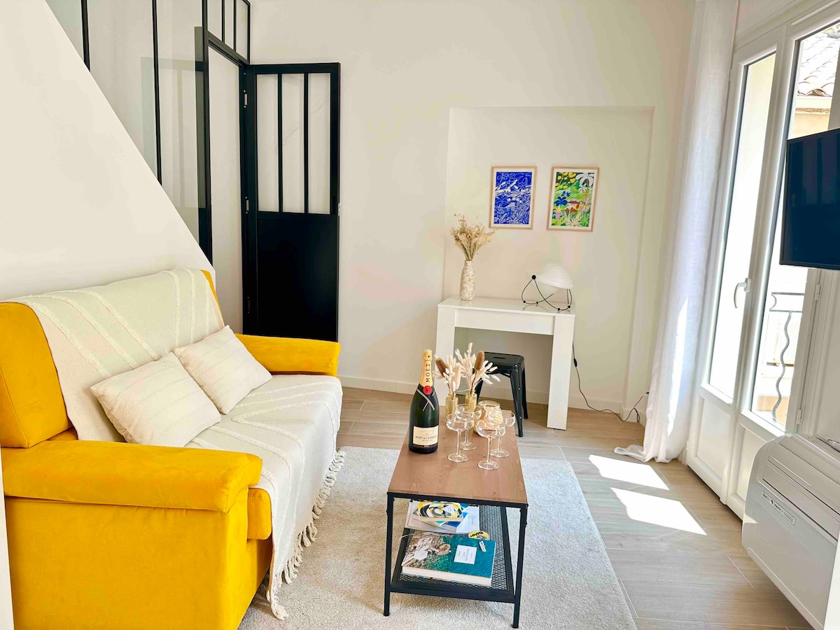 A bright living area features a yellow sofa positioned beside a coffee table, set with glasses and a bottle of sparkling wine. Artwork adorns the wall, complemented by a light-filled corner with large windows and a view of the balcony.