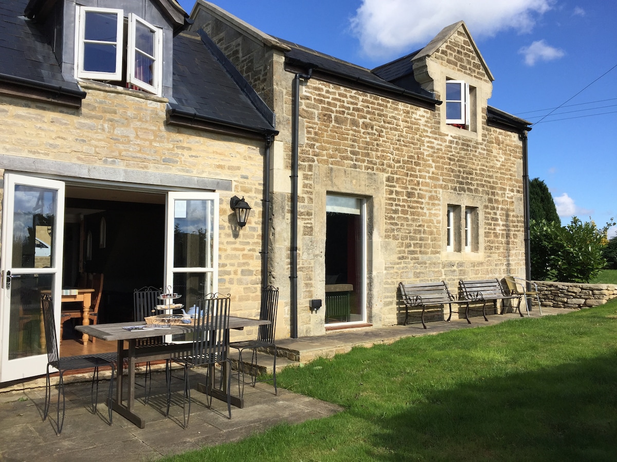 The exterior of the cottage displays a stone façade, complemented by large glass doors leading to a dining area. A spacious patio with a table and multiple chairs is visible, surrounded by well-maintained grass and framed by natural greenery under a clear blue sky.