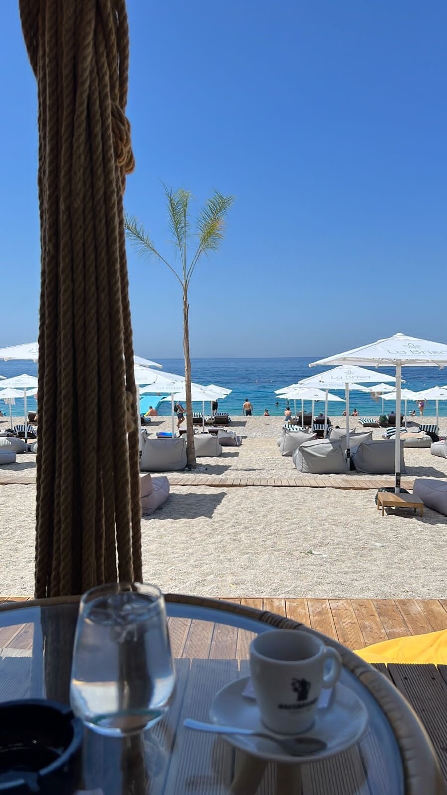 A view from a terrace towards a sandy beach, showcasing numerous sun loungers and white umbrellas. The azure sea extends into the horizon under a clear blue sky. A small table with a glass of water and a coffee cup is positioned in the foreground.