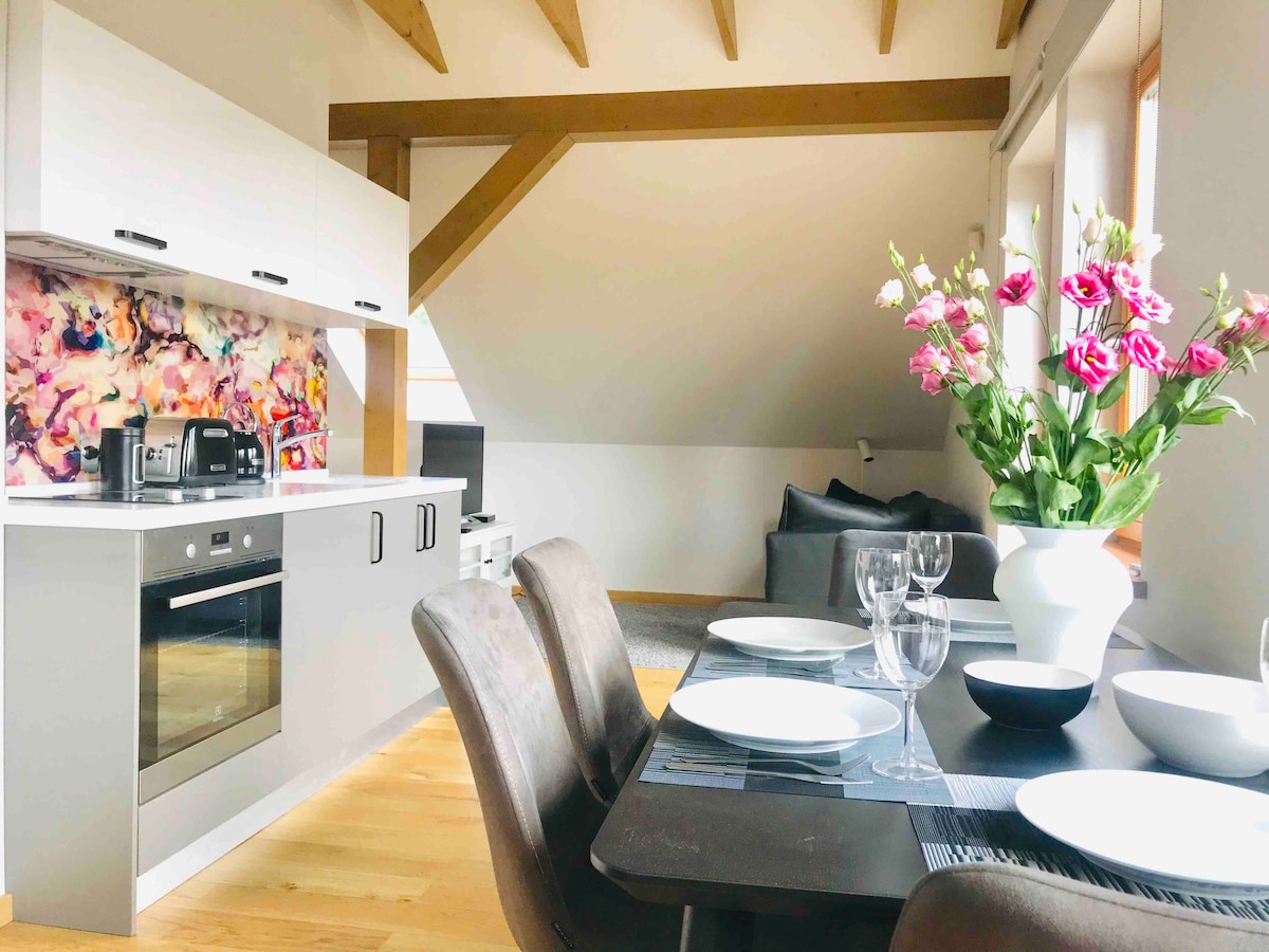 A modern kitchen and dining area is featured with stylish cabinetry and a colorful backsplash. A spacious dining table is set with dishes and glassware. Soft natural light fills the room, highlighting a comfortable couch in the background and exposed wooden beams overhead.
