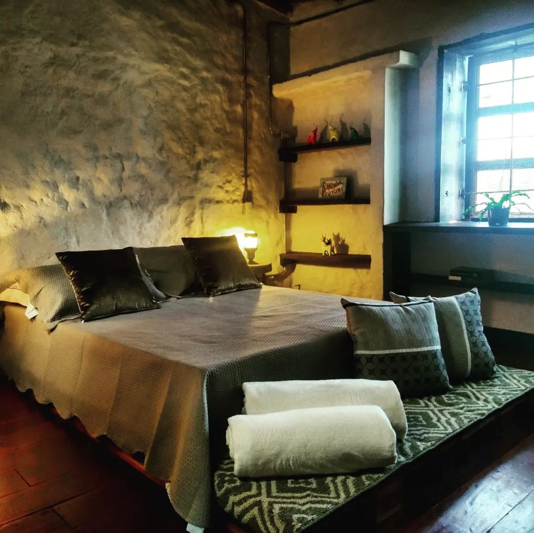 A cozy bedroom features a spacious bed with decorative pillows, complemented by warm lighting from a bedside lamp. A window allows natural light to illuminate the room, while textured walls and a small shelf with decorative items add character to the space.