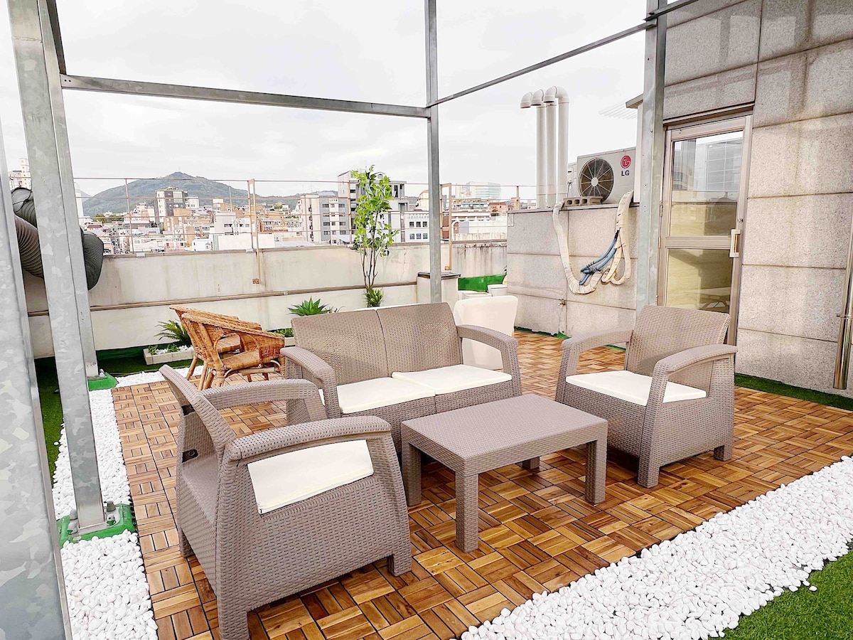 A rooftop garden space is shown, featuring a brown wicker seating arrangement with a sofa and three armchairs surrounding a small table. The area is enhanced by decorative gravel and planters, offering a scenic view of the city skyline.