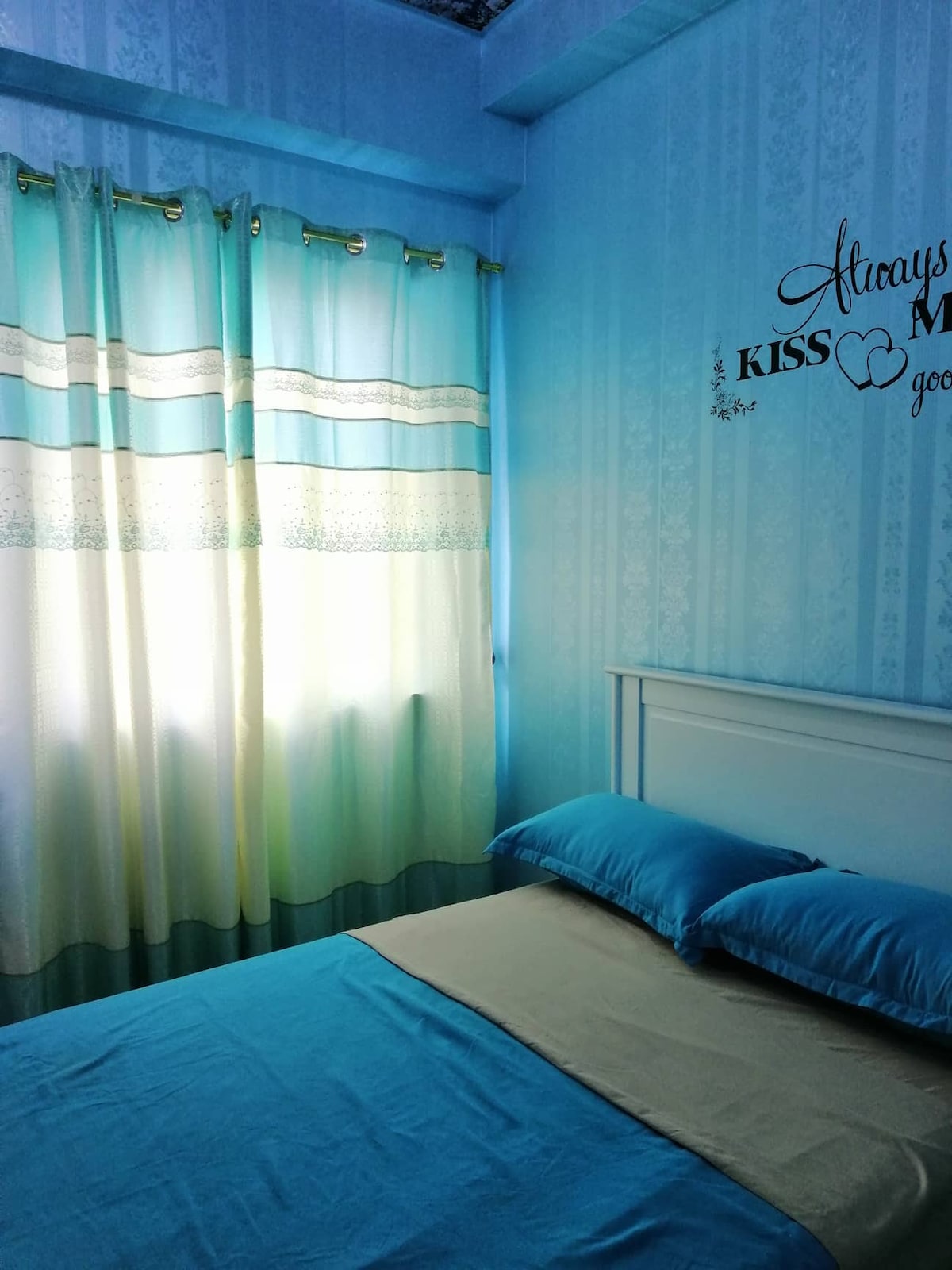 A calming bedroom is presented with soft blue wall accents and light-filtering curtains featuring a gradient design. The small double-size bed is topped with an orthopedic mattress, complemented by accent pillows. Natural light enters through the window, highlighting the serene atmosphere.