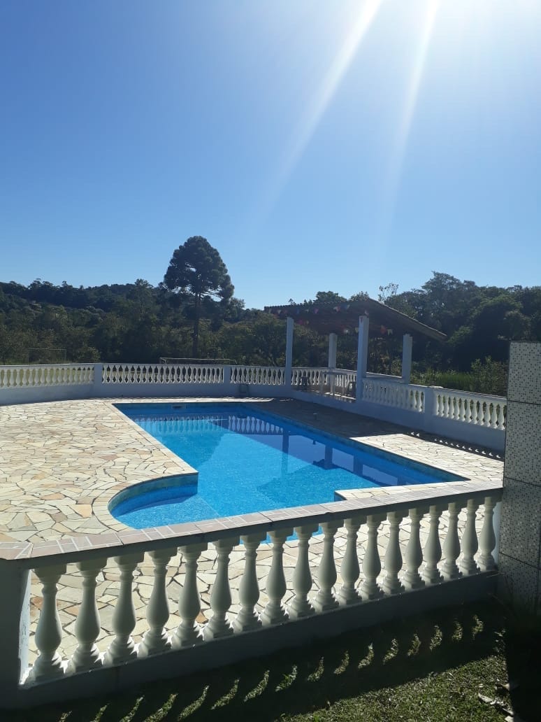 A swimming pool is surrounded by a spacious stone deck, featuring white balustrades. Lush greenery and trees are visible in the background, providing a natural setting. Sunlight reflects off the water, creating a bright and inviting scene.