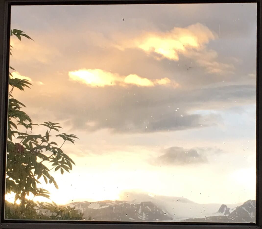 A scenic view is framed through a large window, highlighting soft, pastel-colored clouds illuminated by sunlight. Mountains are visible in the distance, partially shrouded in mist, while the foreground features greenery gently silhouetted against the evening sky.