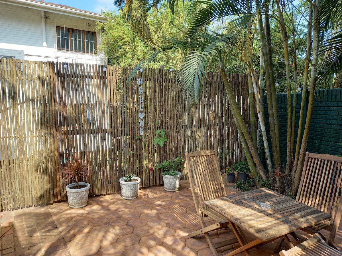 A shaded outdoor area features a wooden table with two chairs surrounded by potted plants. The space is enclosed by a bamboo fence, providing privacy. Lush greenery is visible, enhancing the natural ambiance of the courtyard.